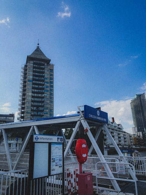 A clear daytime view of a large white suspension bridge spanning a wide river in an urban setting, with modern high-rise buildings and office structures visible in the background. The bridge features