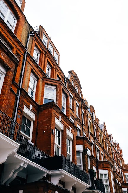 Exterior view of a row of white residential townhouses with blue doors and window frames located on a cobblestone street in Chelsea. The buildings feature small front gardens with decorative plants an